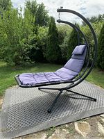 Front outdoor view of Vivere Dream Chair hammock chair with blue cushions on black metal frame, placed on outdoor mat surrounded by trees and grass.