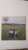 Front cover of America 'View From The Ground' vinyl record album showing band members sitting in a green field with guitar.