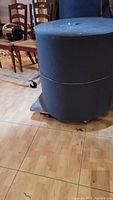 Full view of a large roll of blue acoustic cloth fabric standing upright indoors on a wood floor.