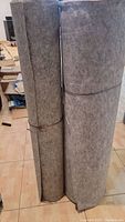 Full view of two large rolls of gray acoustic cloth standing vertically, tied to keep the roll secure. The cloth has a felt-like texture.