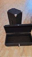 Two rectangular cases covered in acoustic cloth, one open showing hollow interior for audio gear, both with metal corners and hinges