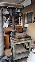 Full view of the vintage Delta drill press mounted on a metal stand with storage shelves, showing wear and rust.