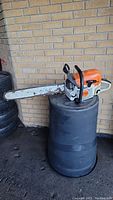 Orange and grey Stihl MS362C chain saw resting on a black plastic drum against a brick wall