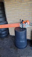 Full view of chain saw resting on barrel, orange scabbard installed on guide bar, brick wall background