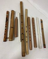 Top-down view of 9 assorted wooden and plastic flutes arranged side by side on white fabric background.
