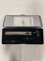 Open view of the black plastic carry case showing the silver cylindrical electric tool, two black attachments, a charger, and instruction manual in molded insert.