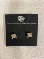 Front view of pair of square gold-coloured earrings studded with clear cubic zirconia stones, presented on branded black card.