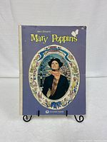 Front cover of Walt Disney's Mary Poppins soft cover movie book showing Mary Poppins portrait with decorative frame on purple background.