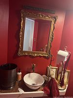 Photo of the bathroom area showing the ornate gold-tone framed mirror, sink bowl, standing toilet paper holder with red rope decoration, brass hand towel holder with burgundy towel, framed prints, and black waste bin.