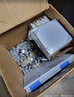 Open box showing assorted French ceramic tiles including decorative square tiles with blue corner designs and gray mosaic tiles along with plain white tiles.