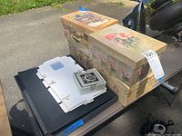 Photo shows two decorative boxes with floral vintage designs; one cloth covered and one cardboard covered, along with a black case and white flat item on the table.