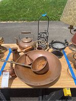 Photo showing large hammered copper bowl with ladle, small copper pitcher, medium kettle, decorative wrought iron wine holder, and pedestal bowl on wooden table outdoors.