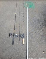 Photo showing two fishing rods with reels and a fishing net with a green net head and aluminum handle laid on the ground.