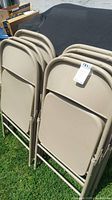 Photo showing 8 beige/tan metal folding chairs stacked together outdoors on grass.