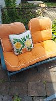 Bench viewed from front with orange cushions and decorative lemon pillow, placed on stone patio.