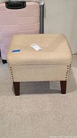 Front angled view of beige woven fabric storage ottoman with nail head trim and wooden legs, showing missing nail head and surface wear.