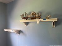 Wide-angle photo of two white wooden wall shelves mounted on a blue wall. One larger shelf holds three miniature wooden sailing ships and a ship in a bottle. The smaller shelf is empty.