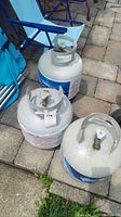 Full view of three propane tanks arranged on outdoor stone patio with a blue lawn chair in background. Tanks are gray metal with blue branding labels and top valve handles.