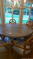 Full view of the large oval dining table with wooden top and integral chairs with patterned cushions, by window with chandelier above.
