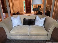 Front view of beige loveseat with three decorative pillows (two white patterned and one dark brown). Shows stains and upholstery details.