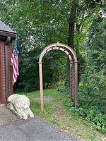 Full view of brown plastic garden arch standing on grass near driveway, adjacent to a house with an American flag and stone lion statue.