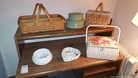 View of multiple baskets including a wooden Longaberger basket with handles and dividers, two pastel keepsake boxes, a white woven sewing basket with floral cushioned lid, and smaller white woven baskets.