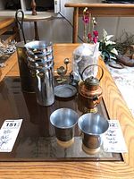 Vintage cups with ribbed bases, copper kettle, brass pitcher, coffee canister, and bird-shaped cutter rest on a table