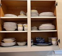 Two cabinet shelves filled with plates, bowls, and cups in mixed sets, showing variety of designs and materials.