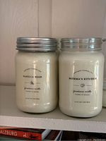 Two farmhouse candles in glass jars: Vanilla Bean and Momma's Kitchen with metal lids on top and label details visible.