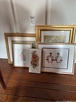 Four framed art prints leaning against a white textured wall on a wooden floor, showing varied sizes and frames.