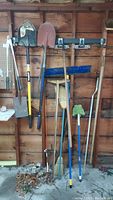 Full view of garden tools hanging on garage wall including shovels, rakes, brooms, pruners.