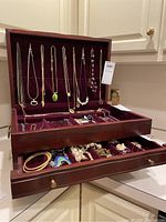 Top open section of jewelry box showing necklaces hanging and velvet compartments with rings and small jewelry pieces.