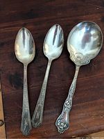 Three silver plated spoons shown on wooden surface including two simpler spoons and one ornate spoon