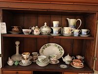 Wide view of multiple tea cups, saucers and teapots displayed on wooden shelf, showing variety and condition of the set