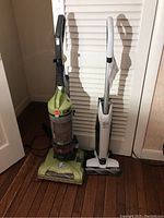 Two upright vacuum cleaners side by side: green Hoover WindTunnel and white Hoover Onepwr cordless stick vacuum.