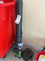 Battery operated leaf blower with black and green design, standing upright next to a red plastic bin, showing blower tube and handle.