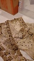 Sheer beige fabric with rich gold and bronze embroidery shown beside plastic storage bin and cardboard box
