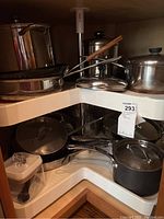 Corner kitchen cabinet with multiple pots and pans, mostly stainless steel with lids and a cast iron skillet in front.