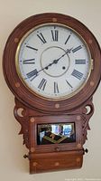Front view of regulator clock showing round white clock face with Roman numerals, wooden bezel with circular brass inlays.