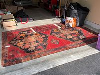 Full view of large 9'x6' rug with red base, black and orange geometric and floral motifs, placed partially in garage.