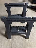 Two black plastic folding saw horses standing on concrete floor with hooks on the sides and cross bars at the top.
