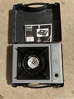Portable Coleman single burner butane stove inside black plastic carrying case, with manual visible.