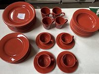 Photo showing stacked terracotta dinner plates, sandwich plates, soup bowls, 4 coffee mugs, and 4 coffee cups with saucers arranged on table.