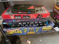 Photo of five boxed items including a green metal poker set on top, Home Alone 2 board game, Batman Forever board game, and Teenage Mutant Ninja Turtles Electric Pinball board game boxes stacked on a shelf.
