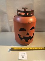 Front view of orange metal pumpkin lantern with jack-o-lantern face cut out and measuring tape for scale