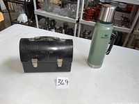 Photo showing the black Thermos lunch box and green Stanley thermos side by side on a white surface with shelves of various items in the background.