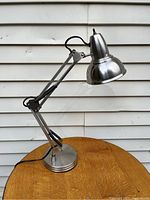 Full view of the metal desk task lamp with retro design on round wooden surface against a white siding background. Shows articulated arm and base.