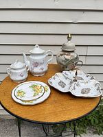Photo displaying all five china items on a round wood table outside, including teapot, sugar jar, covered jar, divided pastry plate, and floral plate.