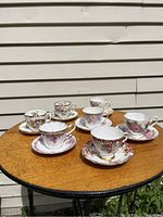 Full view of 7 bone china teacups and their matching saucers laid out on a wooden table, showing various floral patterns and gold trim.