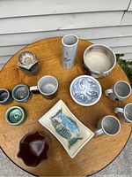 Overview of multiple studio pottery items arranged on a round wooden table. Includes mugs, decorative trays, bowls, and vases with varied glazes.
